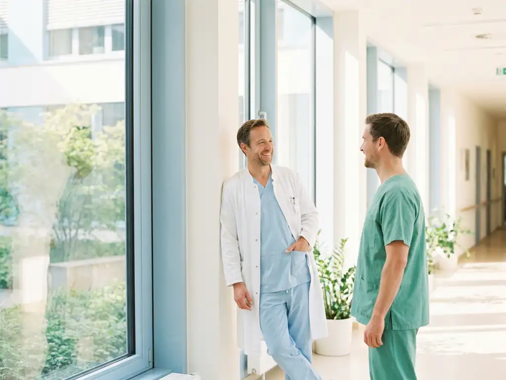facharzt-krankenhaus-kollegen-gespraech.jpg Lächelnder Facharzt im weißen Kittel unterhält sich mit einem Kollegen in einem hellen deutschen Krankenhausflur.